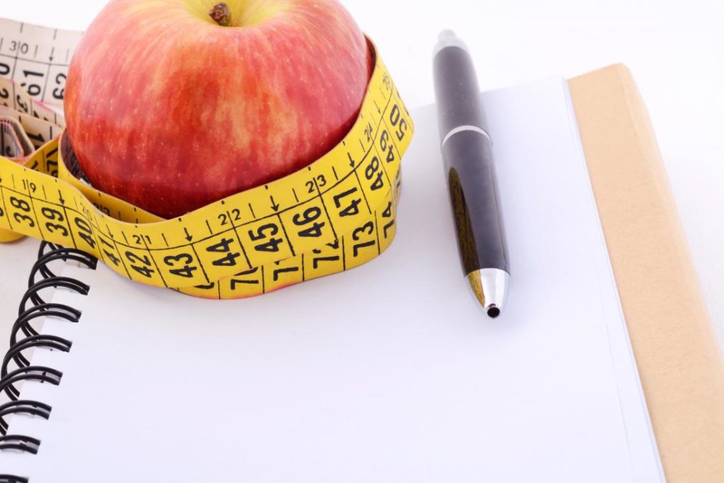 Planning an intermittent fast: A measuring tape wrapped around an apple resting on a notepad with a pen next to it. 